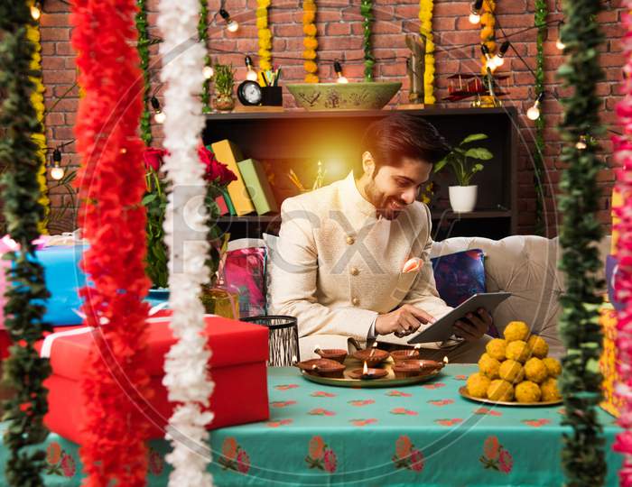 Indian man using tablet touchscreen computer on festival day while wearing traditional outfit. sitting on sofa with gifts and sw