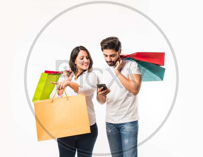 Indian young couple shopping bags and smart phone or mobile, clicking selfie or locating store, standing isolated over white bac