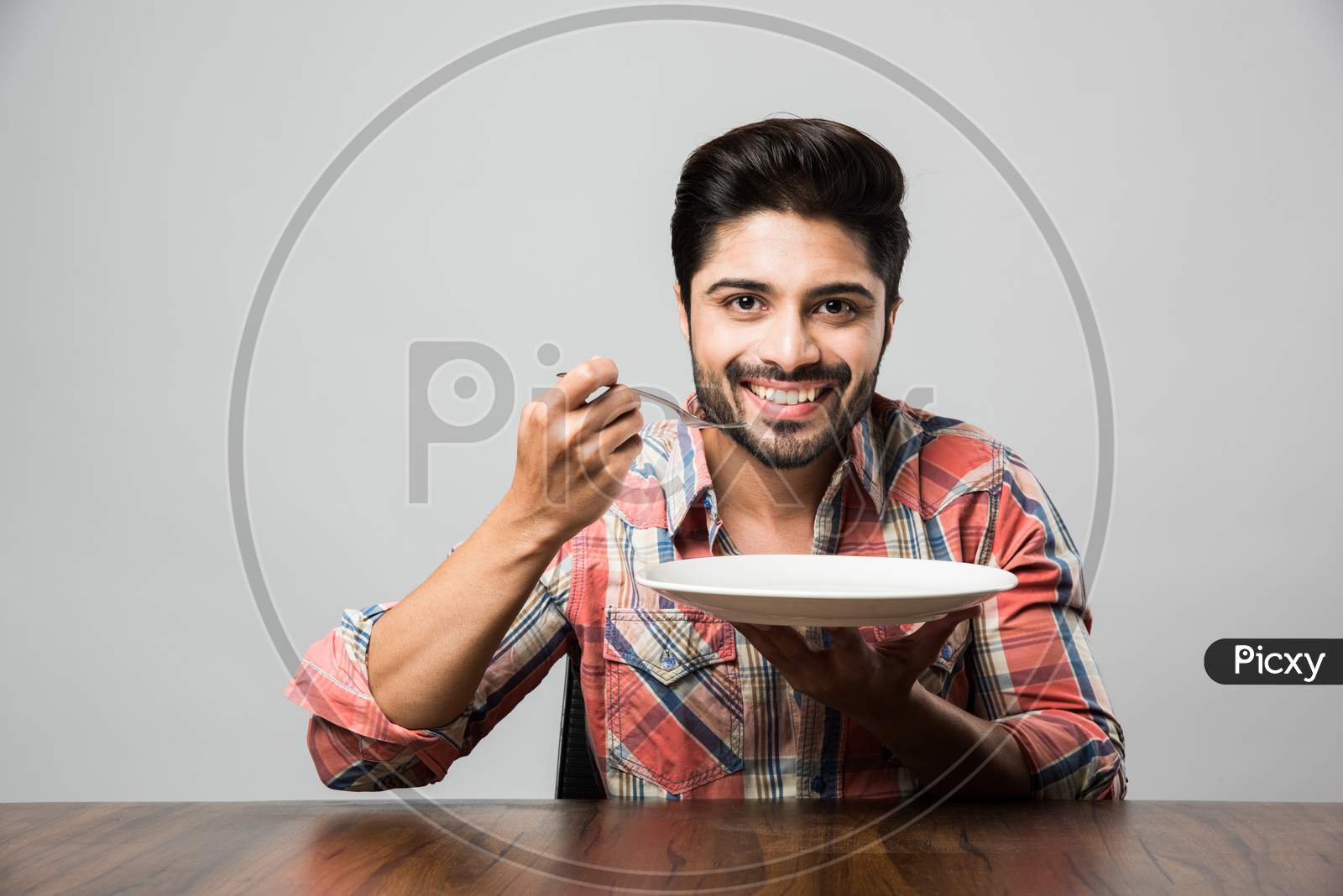 empty plate and Indian man with beard holding spoon and fork, wearing checkered shirt and sitting at table