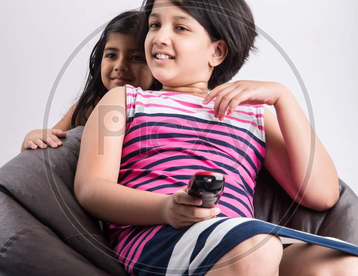 Indian small girls / sisters watching Television / TV while holding remote control, sitting over bean bag