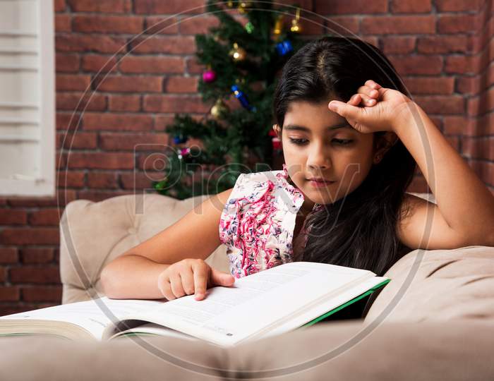 Cute little Indian/asian girl reading book while sitting sofa or couch at home