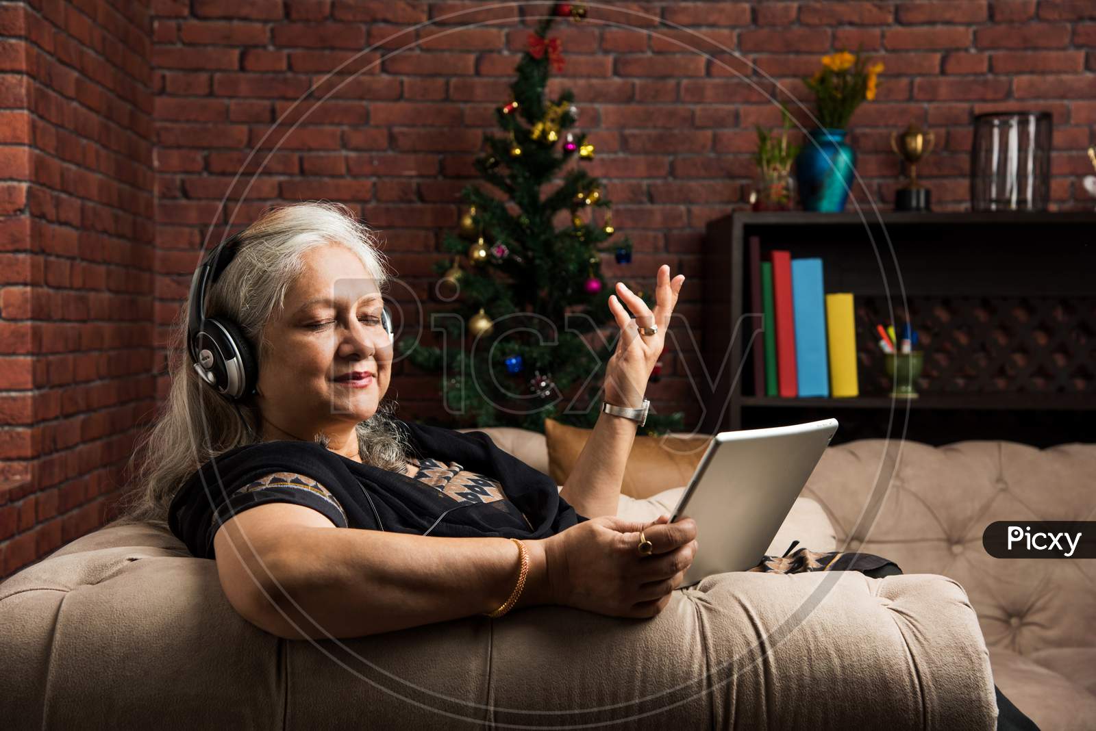 senior Indian/asian women listening to music using headphones while holding a tablet computer and sitting on sofa or couch at ho
