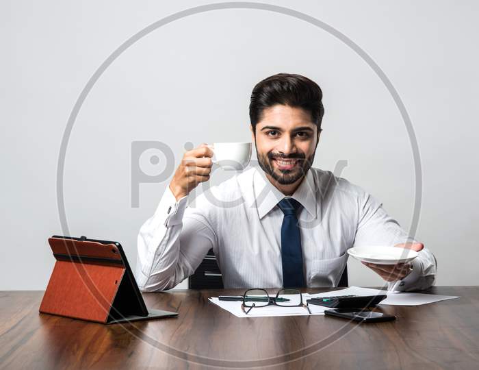 Indian businessman having coffee / tea at work / office, sitting at desk or table holding cup and saucer