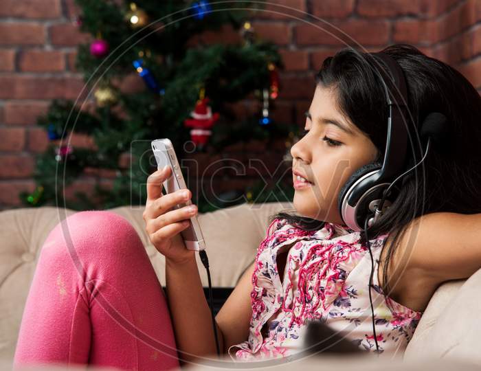cute little indian/asian small girl listening to music using headphones or earphone on smartphone, sitting on couch or sofa at h