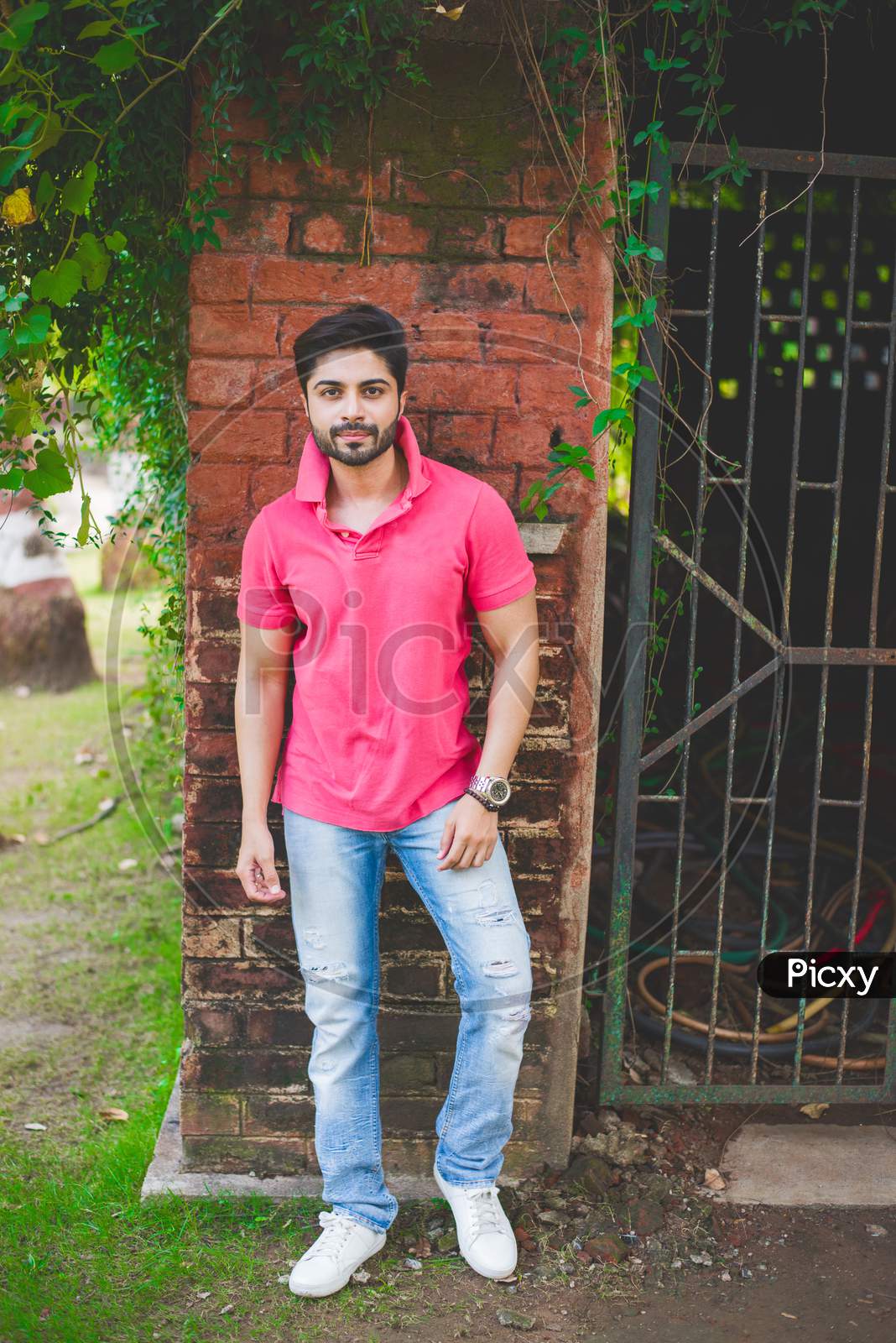 Bearded Indian young man outdoor, standing against red brick wall at garden / park
