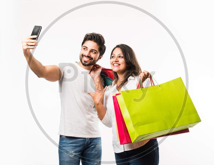 Indian young couple shopping bags and smart phone or mobile, clicking selfie or locating store, standing isolated over white bac