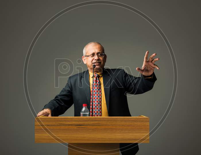 Happy Indian/asian handsome senior businessman speaking with mic at podium in office or auditorium presenting something
