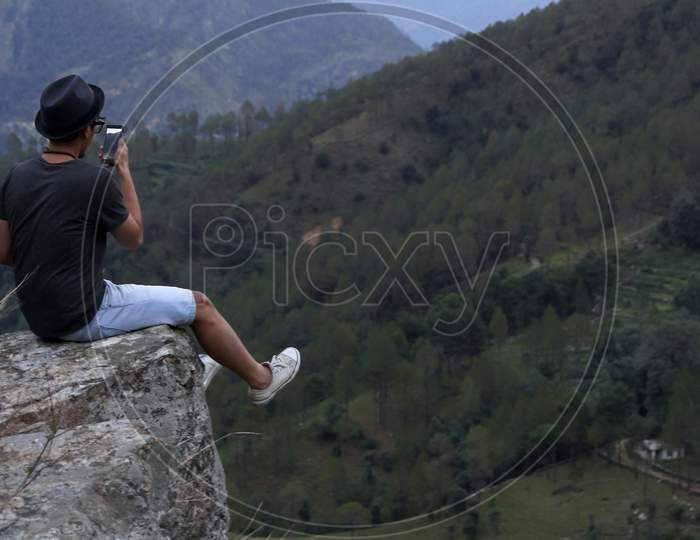 Young Man Wearing A Cool Hat Taking Photos On His Cellphone Of A Mountain Valley While Sitting On The Edge Of A Rock.