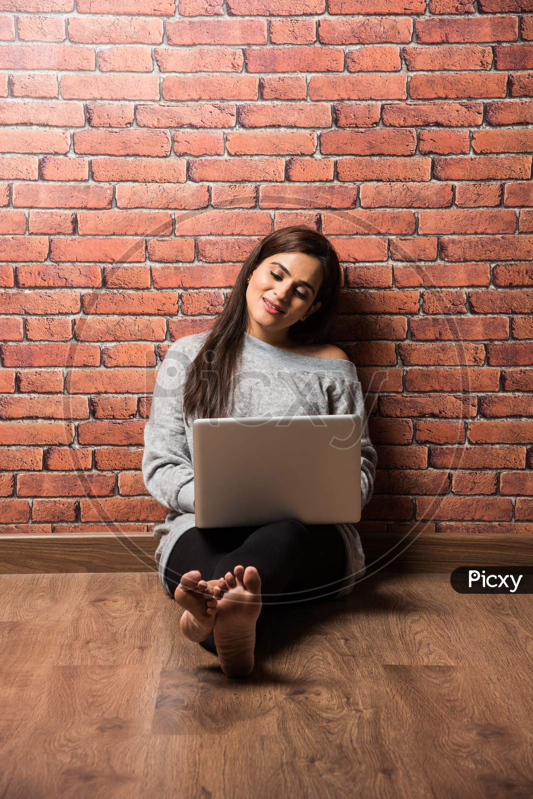 Indian girl with laptop sitting over wooden floor against red brick wall
