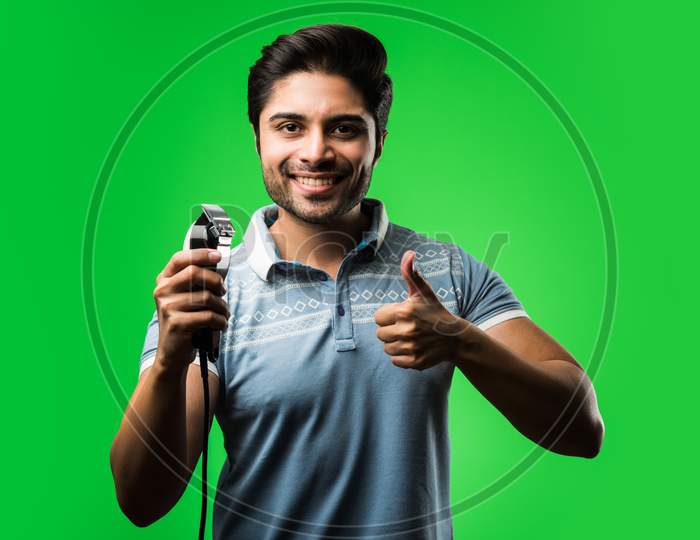 Indian / asian man shaving beard with Electric trimmer or clipper standing isolated over green background