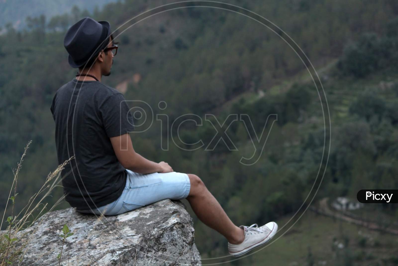 Young Man Wearing A Cool Hat Sitting On The Edge Of A Rock If Front Of A Beautiful Mountain Valley.