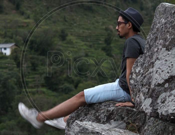 Young Man Wearing A Cool Hat And Waving His Legs While Sitting On The Edge Of A Rock In The Mountains.