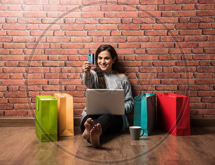 Indian girl shopping with debit / Credit card and laptop while sitting over wooden floor against red brick wall