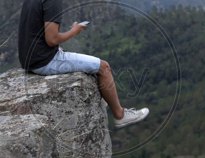 Young Man Wearing A Cool Hat Using His Cellphone While Sitting On The Edge Of A Rock In Front Of A Beautiful Mountain Valley.