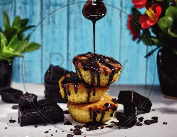 Chocolate syrup is falling on the cupcakes from the top and the chocolate cubes are also there infront of a sky textured background.