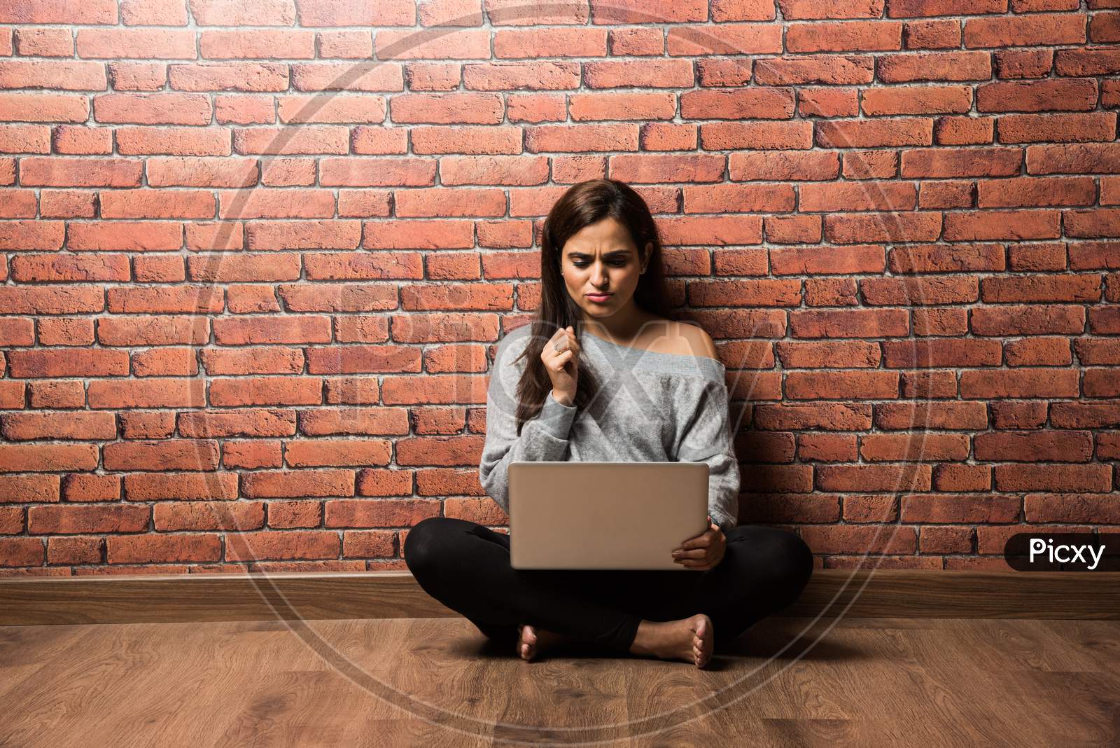 Indian girl with laptop sitting over wooden floor against red brick wall