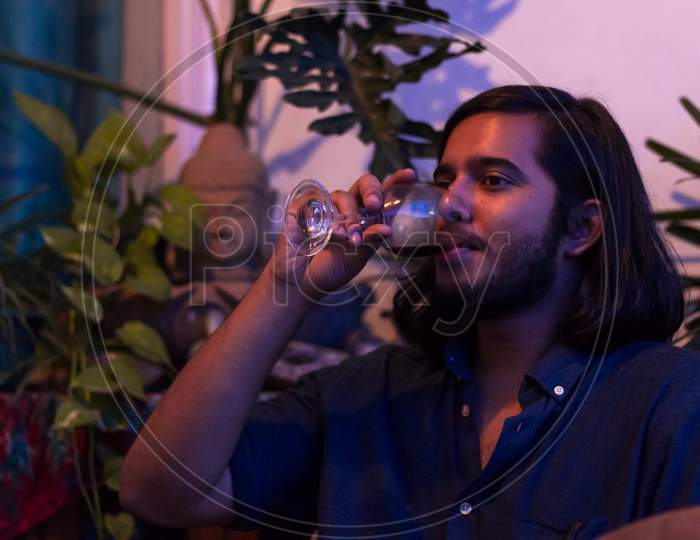 Young Man Drinking Red Wine At Home With Indoor Plants In Background.