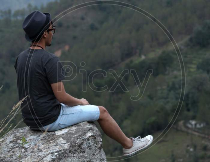 Young Man Wearing A Cool Hat Sitting On The Edge Of A Rock If Front Of A Beautiful Mountain Valley.
