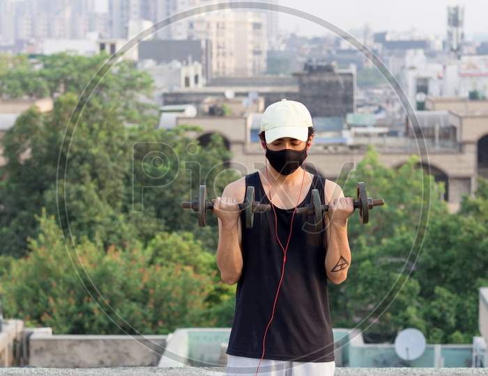 Young Man Avoiding Going To Gym To Maintain Social Distancing And Lifting Dumbbells At His Terrace While Wearing A Mask To Protect Himself From Corona Virus Infection During World Pandemic.