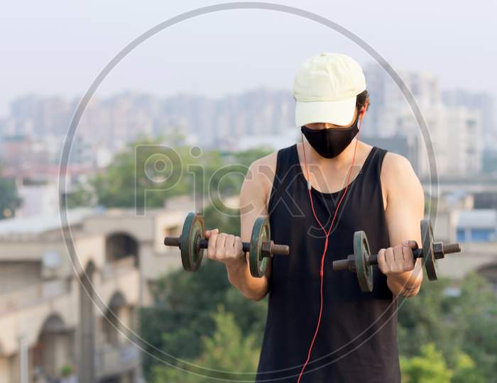 Young Man Avoiding Going To Gym To Maintain Social Distancing And Lifting Dumbbells At His Terrace While Wearing A Mask To Protect Himself From Corona Virus Infection During World Pandemic.