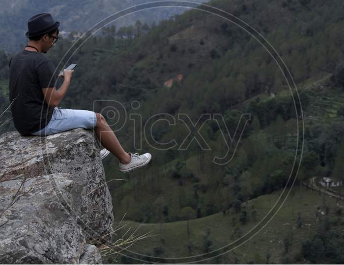 Young Man Wearing A Cool Hat Using His Cellphone While Sitting On The Edge Of A Rock In Front Of A Beautiful Mountain Valley.