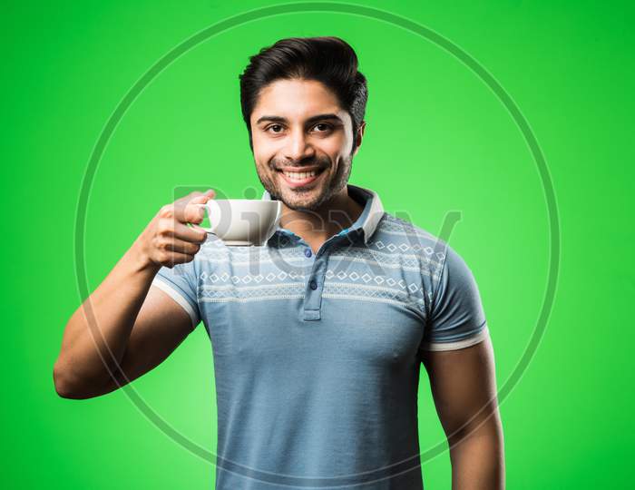 Indian man with tea / coffee cup or mug. Drinking, presenting or holding while standing isolated over green background