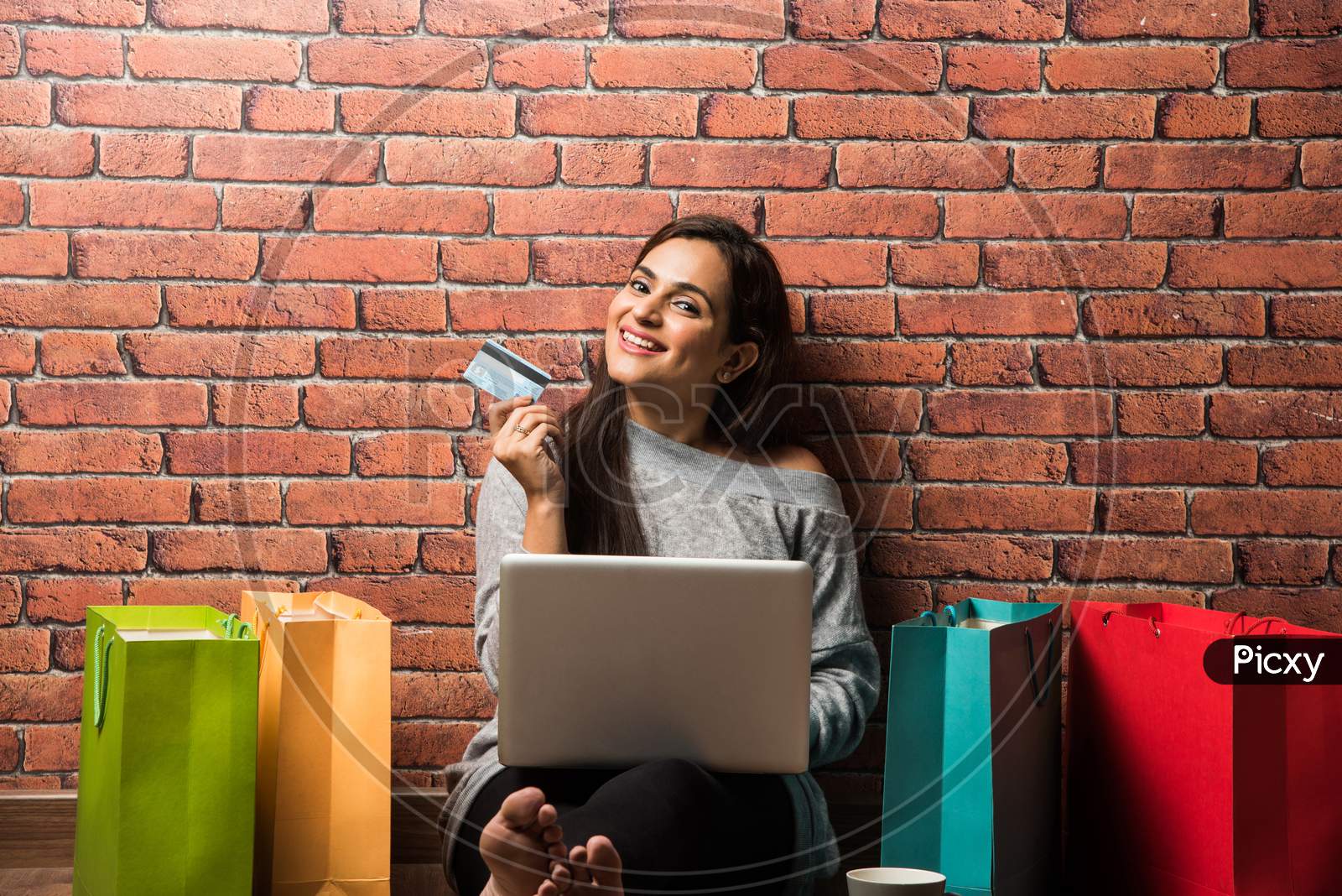 Indian girl shopping with debit / Credit card and laptop while sitting over wooden floor against red brick wall