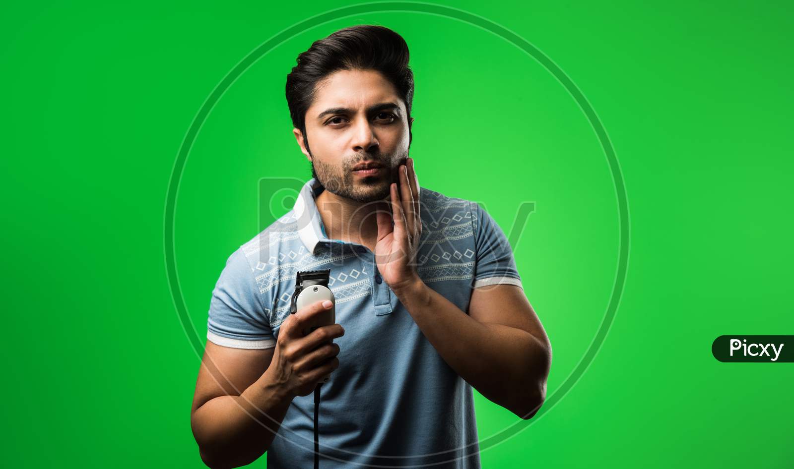 Indian / asian man shaving beard with Electric trimmer or clipper standing isolated over green background