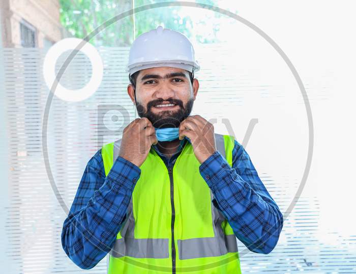 Young Engineer Taking Off Mask Smiling, Closeup Of Beard Man Wearing Blue Shirt With Yellow Vest And White Helmet, Back To Work After Lockdown Ends Due To Covid-19 Pandemic, New Normal