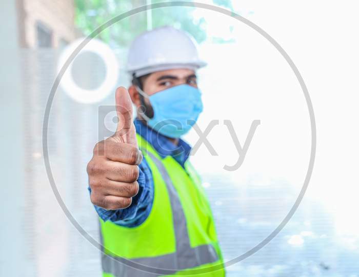 Young Engineer Wearing Take Off Mask Thumbs Up Gesture With Hand, Approving Expression, Man Wearing Blue Shirt With Yellow Vest And White Helmet, Selective Focus On Hand, Covid-19 Pandemic