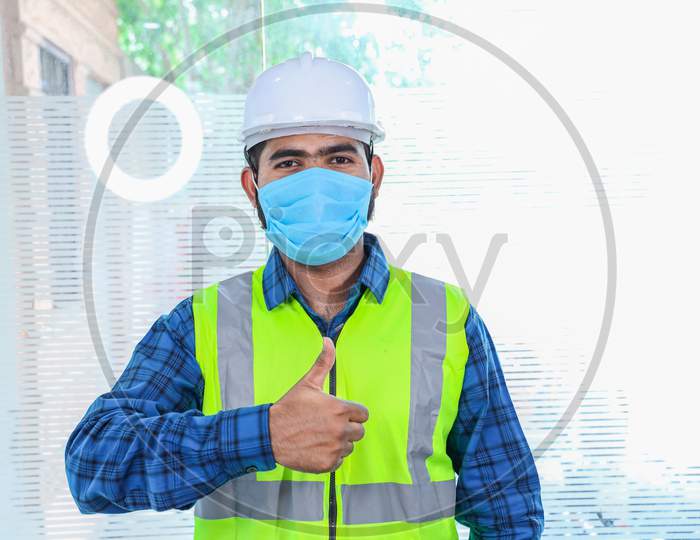 Young Engineer Wearing Mask Thumbs Up Gesture With Hand, Approving Expression, Man Wearing Blue Shirt With Yellow Vest And White Helmet, Back To Work After Lockdown Ends Due To Covid-19