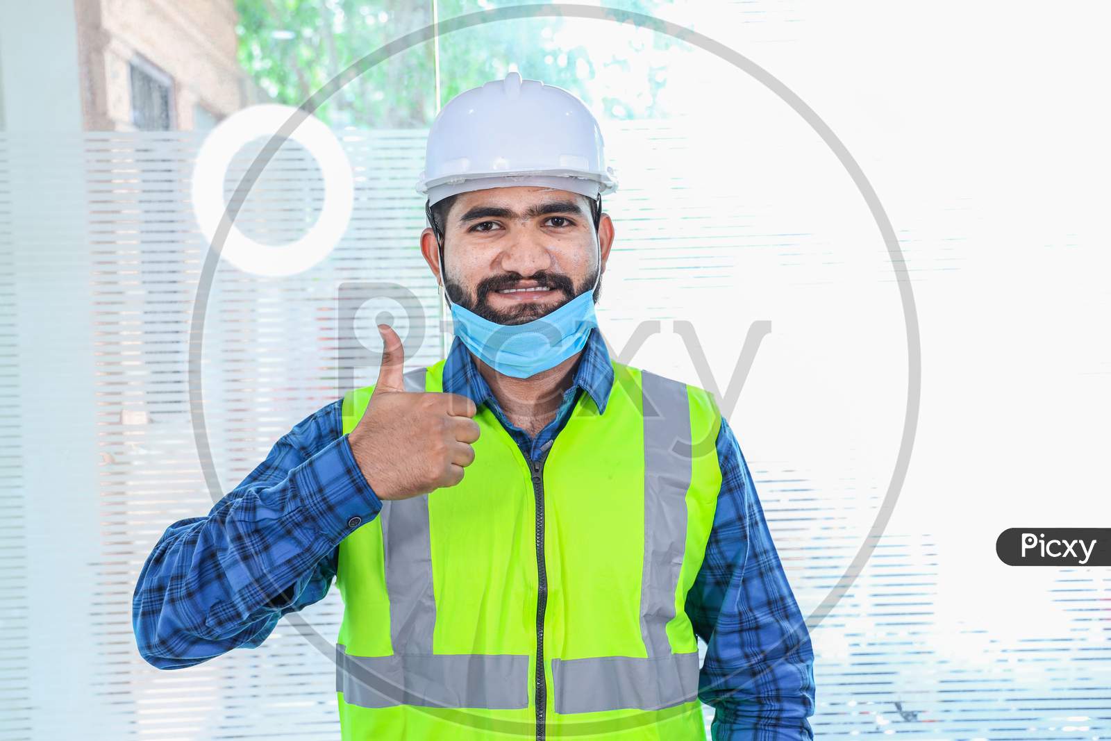 Young Engineer Wearing Take Off Mask Thumbs Up Gesture With Hand, Approving Expression, Man Wearing Blue Shirt With Yellow Vest And White Helmet, Back To Work After Lockdown Ends Due To Covid-19