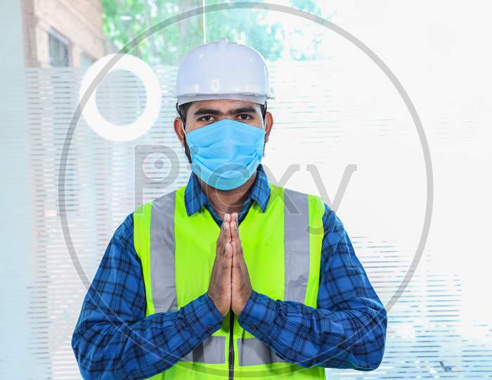 Young Engineer Wearing Mask Doing Namaste, Closeup Of Beard Man Wearing Blue Shirt With Yellow Vest And White Helmet, Back To Work After Lockdown Ends Due To Covid-19 Pandemic, New Normal Lifestyle
