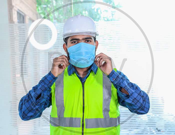 Young Engineer Wearing Adjusting Mask Smiling, Closeup Of Beard Man Wearing Blue Shirt With Yellow Vest And White Helmet, Back To Work After Lockdown Ends Due To Covid-19 Pandemic, New Normal