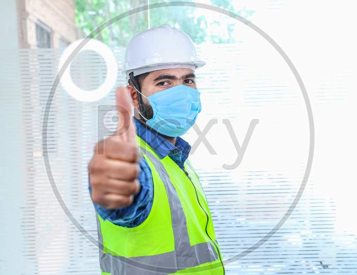 Young Engineer Wearing Take Off Mask Thumbs Up Gesture With Hand, Approving Expression, Man Wearing Blue Shirt With Yellow Vest And White Helmet, Back To Work After Lockdown Ends Due To Covid-19