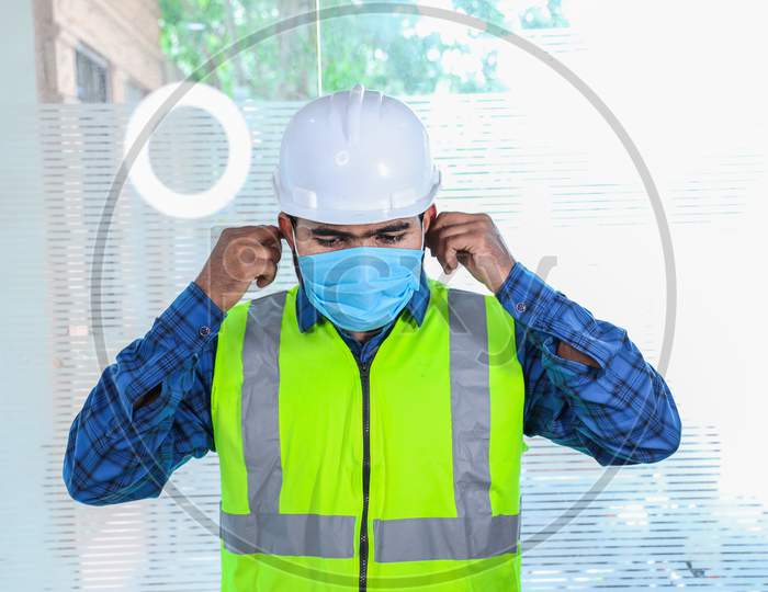 Young Engineer Wearing Adjusting Mask Smiling, Closeup Of Beard Man Wearing Blue Shirt With Yellow Vest And White Helmet, Back To Work After Lockdown Ends Due To Covid-19 Pandemic, New Normal