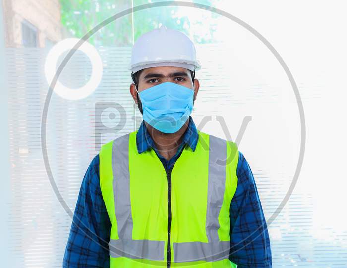 Young Engineer Wearing Mask, Closeup Of Beard Man Wearing Blue Shirt With Yellow Vest And Ywhite Helmet, Back To Work After Lockdown Ends Due To Covid-19 Pandemic, New Normal Lifestyle