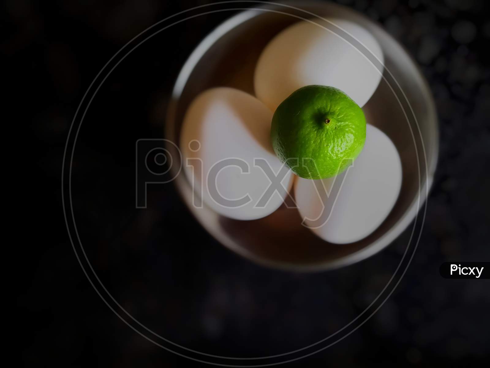 A green lemon on top of three easter eggs under bowl in dark background