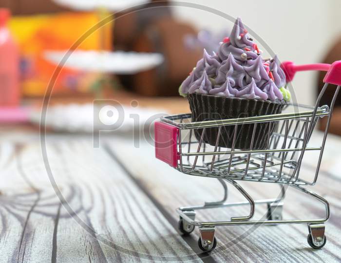 Beautifully Iced Cupcake With Purple Frosting And Black Chocolate Base On A Shopping Cart On A Wooden Floor With Blurred Out Background