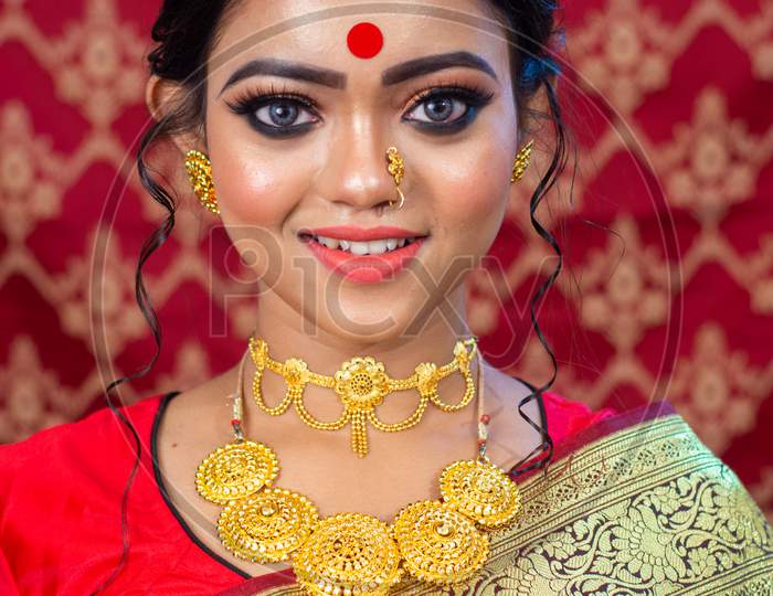 Stunning Indian bride dressed in Hindu red traditional wedding clothes sari embroidered with gold jewelry and a veil smiles tender