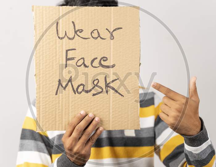 Young Man Holding A Cardboard Sign By Covering His Face, Saying Wear Face Mask Message On Isolated Background To Protect From Coronavirus Or Covid-19.