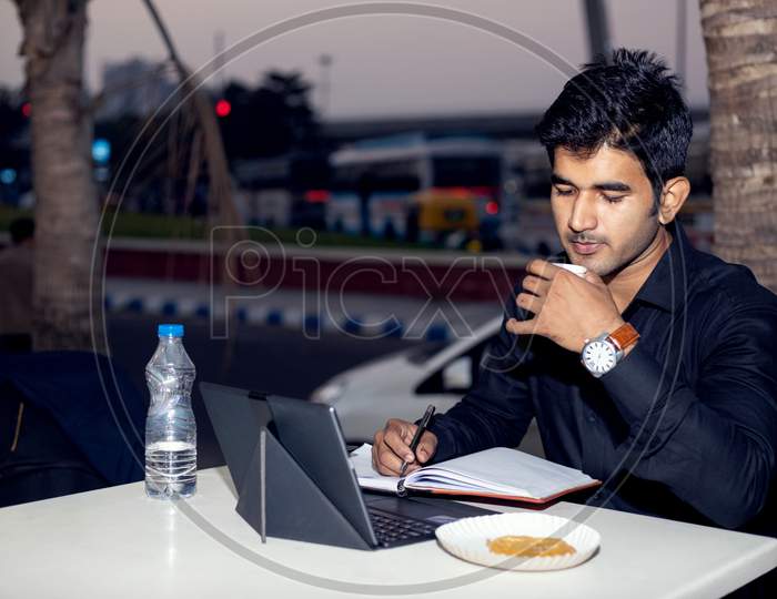 Young Indian man working on laptop from outdoor cafe center while eating snacks and coffee.