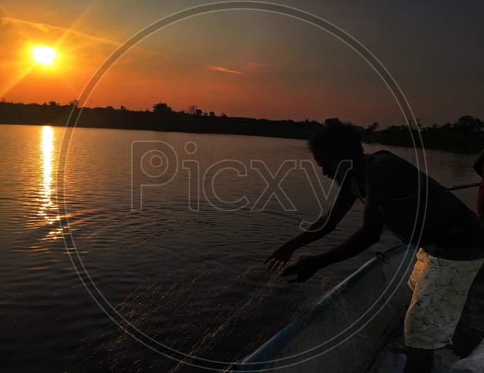 A Man In The Lake At Sunset While His Catching Fishes With Net