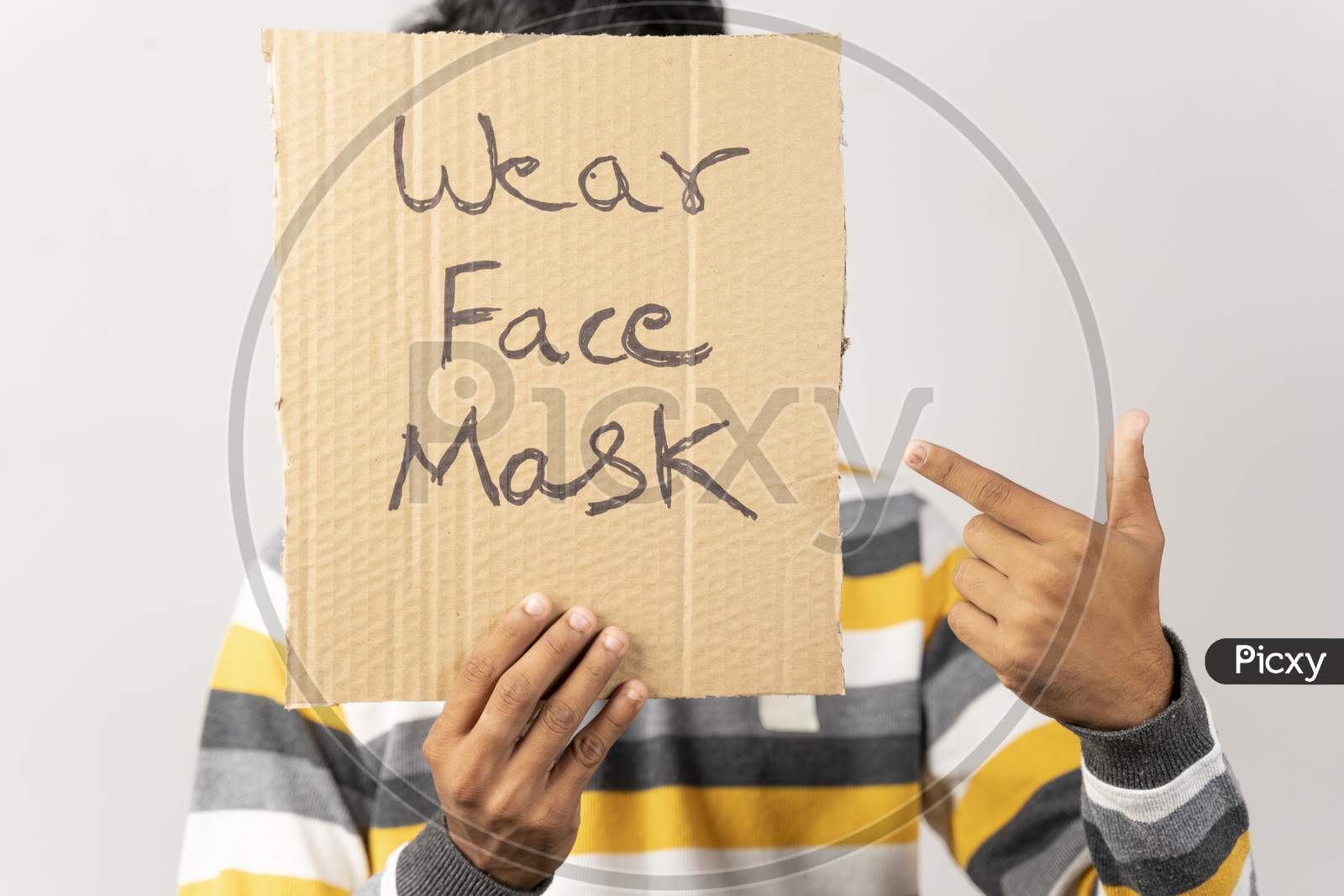 Young Man Holding A Cardboard Sign By Covering His Face, Saying Wear Face Mask Message On Isolated Background To Protect From Coronavirus Or Covid-19.