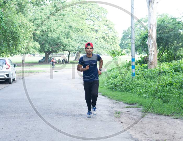 Athletic Indian man running in park. Asian Runner  wearing blue sportswear and red cap jogging outdoors with tree's in background.