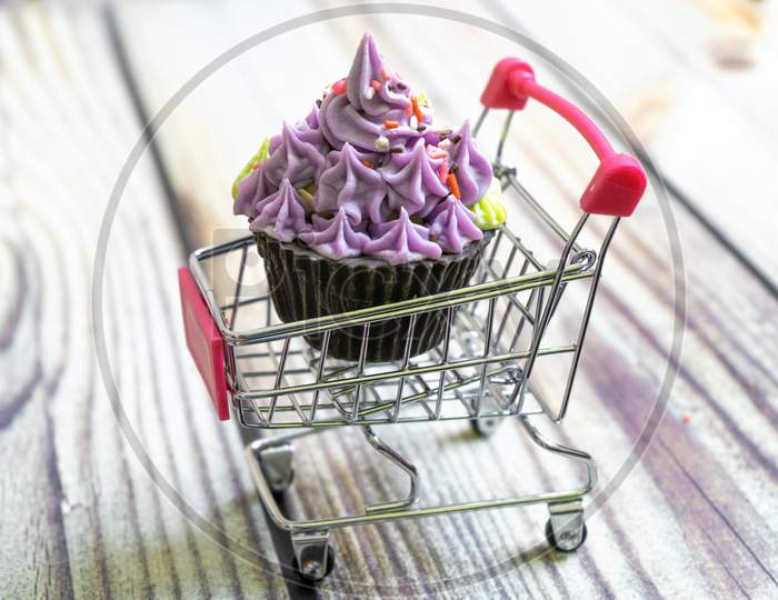 Beautifully Iced Cupcake With Purple Frosting And Black Chocolate Base On A Shopping Cart On A Wooden Floor With Blurred Out Background