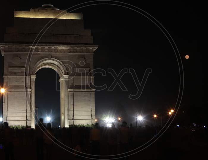 Delhi India Gate At Night With A Red Moon (On The Right) In Sky.