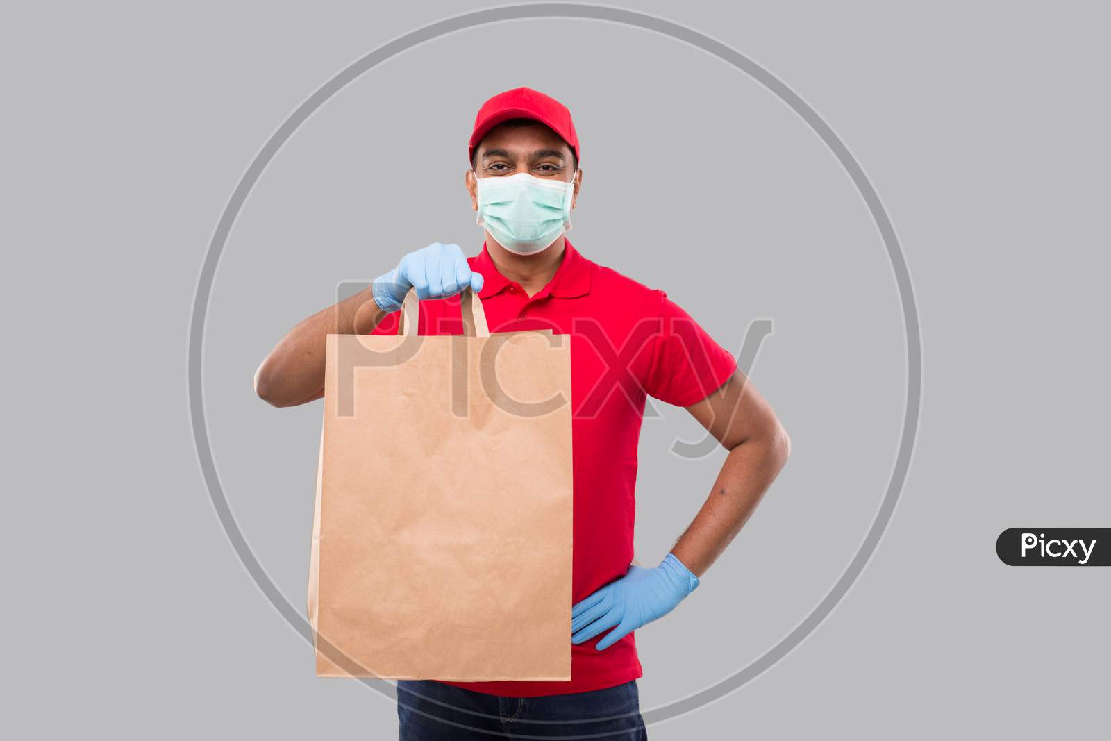 Delivery Man With Paper Bag In Hands Wearing Medical Mask And Gloves Isolated. Red Uniform Indian Delivery Boy. Home Food Delivery. Paper Bag