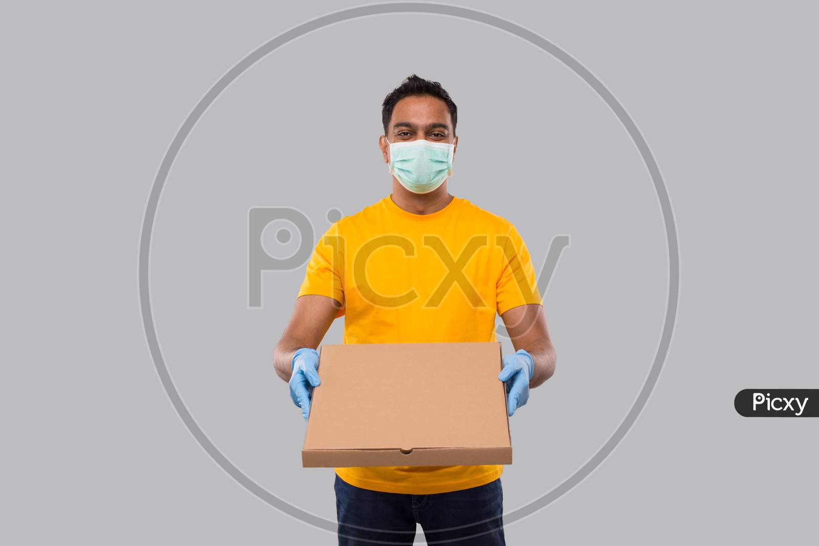 Delivery Man Pizza Box In Hands Wearing Medical Mask And Gloves Isolated. Yellow Tshirt Indian Delivery Boy. Man With Pizza In Hands