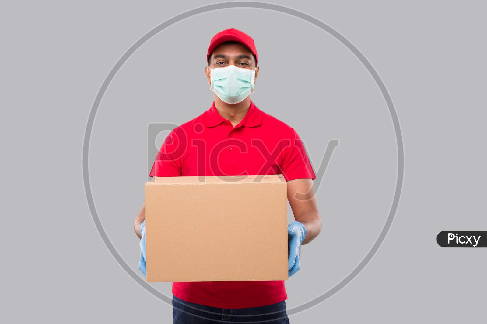 Delivery Man Holding Box In Hands Wearing Medical Mask And Gloves Isolated. Red Tshirt Indian Delivery Boy Watching Side. Home Delivery. Quarantine Hero.